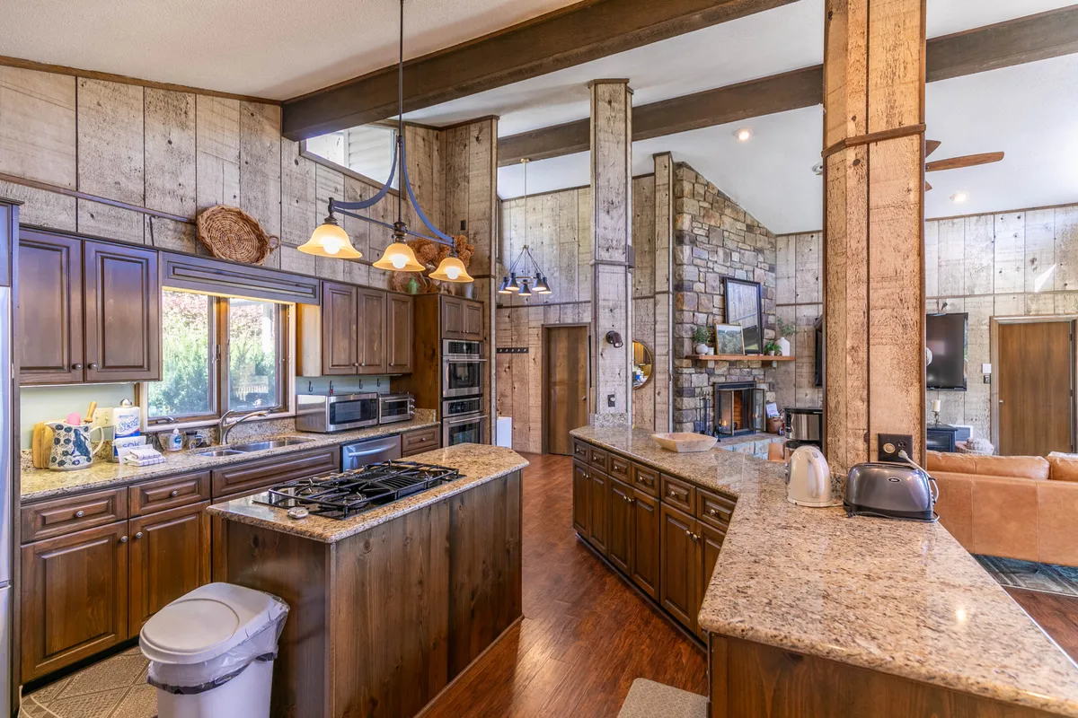 A view from the kitchen area reveals granite countertops and wooden cabinetry, with the stone fireplace and large windows visible in the adjoining living space. The arrangement demonstrates how cooking, dining, and relaxing blend seamlessly in the hub of the home.
