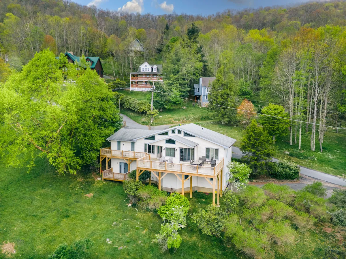 A large front porch framed by surrounding greenery with neighboring houses visible in the background. The porch is designed for family gatherings or outdoor dining, and the setting is perfect for sunrise views or evening stargazing.
