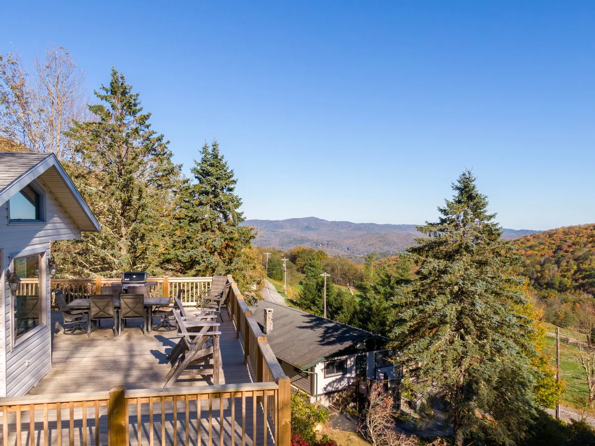 The hero image shows a spacious deck with comfortable seating overlooking a stunning panorama of Blue Ridge Mountains. The deck is surrounded by lush trees, inviting guests to relax, dine outdoors, and soak in the tranquil atmosphere as the sun sets over the peaks.