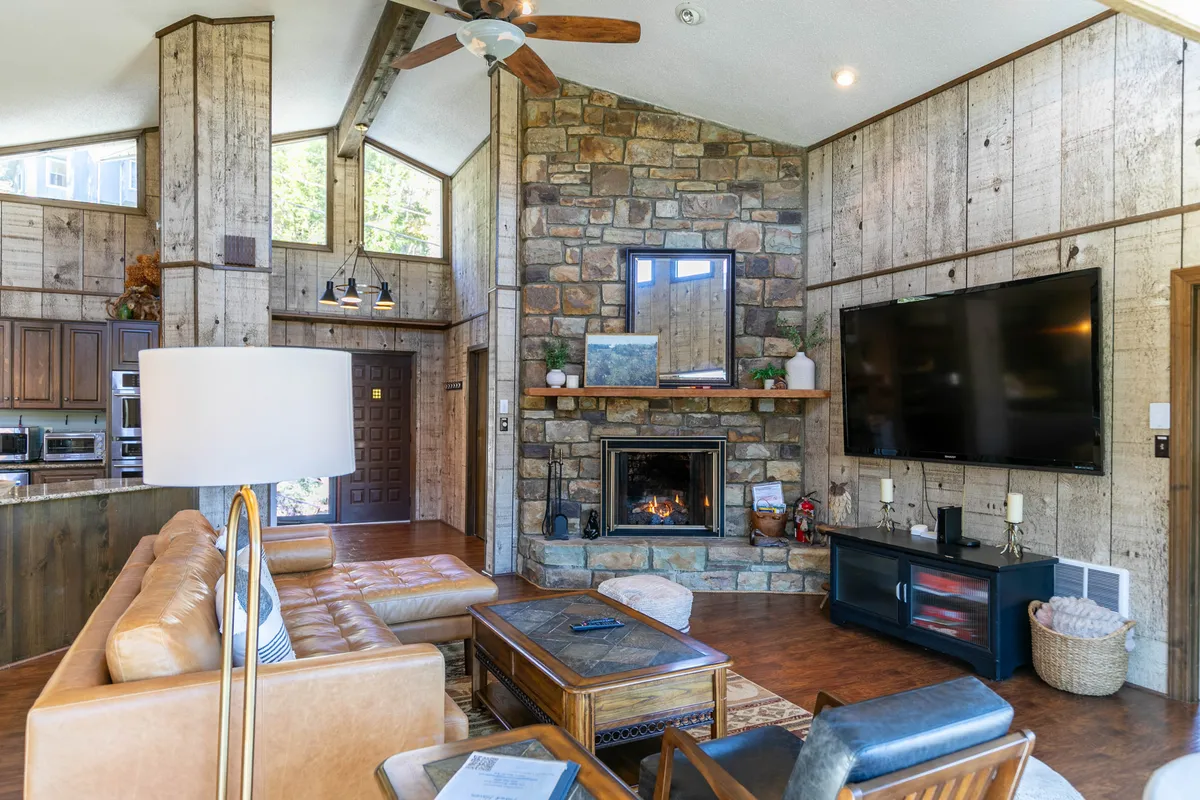 This interior image highlights a living area centered around a stone fireplace, with a leather sofa, large windows letting in ample natural light, and a wooden coffee table. The scene evokes a sense of warmth and coziness, ideal for relaxing evenings after mountain adventures.