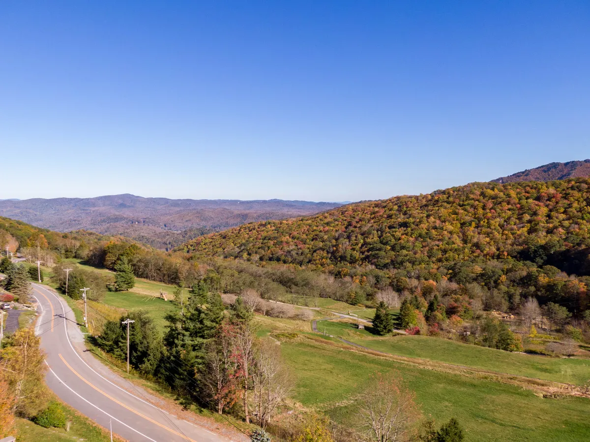 A serene mountain range stretches along the horizon, dotted with autumn foliage and crossed by a winding road. The expansive landscape is bathed in soft sunlight, capturing the area's peaceful charm and offering a glimpse of the region's many hiking and exploration opportunities.