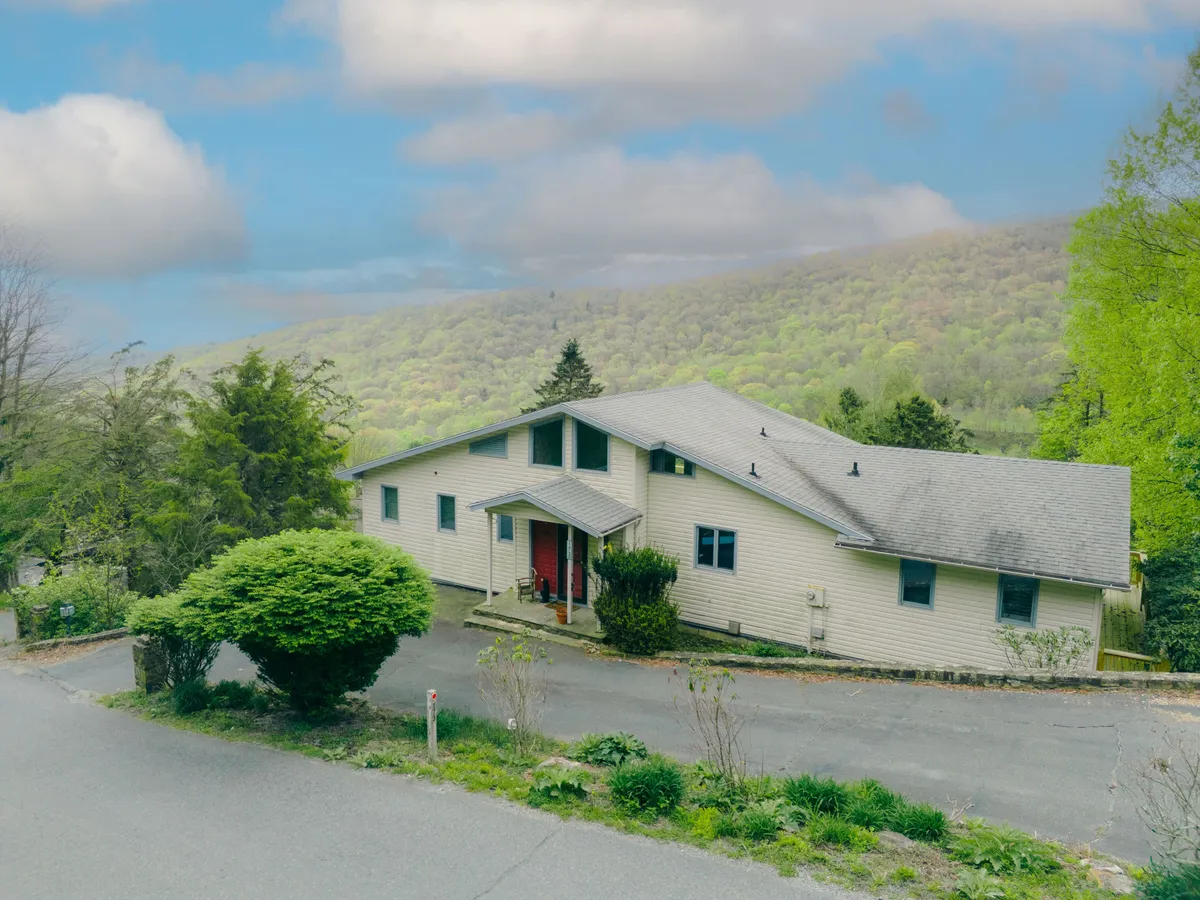 An exterior shot of the house with a view of the paved driveway and the surrounding mountain greenery. Ample parking space is visible, and the tranquil setting hints at the adventure and seasonal challenges of the location.