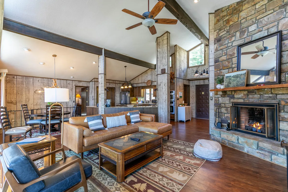 A wide view of the great room features a leather sofa positioned before a striking stone fireplace, with the dining area and a glimpse of the kitchen visible beyond. Rustic wooden beams and paneling create a warm, inviting atmosphere, while large windows fill the space with natural light.