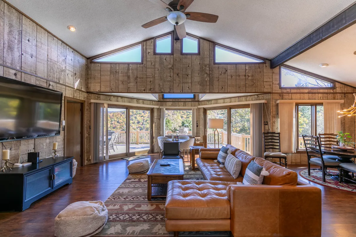 A leather sectional sofa anchors the spacious living area, framed by large windows that showcase picturesque mountain views. The rustic wood walls and ceiling beams enhance the welcoming, mountain-lodge feel. The dining area seamlessly connects to the great room, reinforcing the open-concept design.