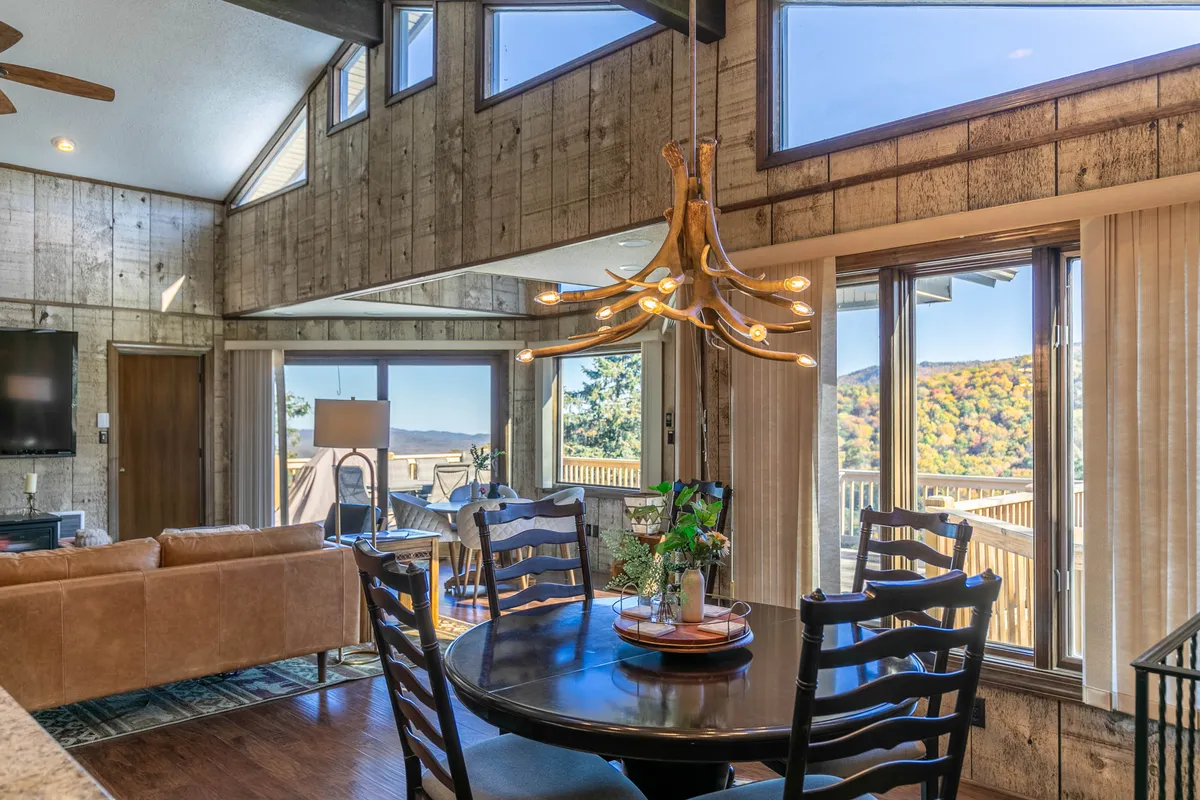 A warm, welcoming interior scene with a dining table positioned near large windows, a chandelier overhead, and a comfortable sofa. Through the windows, natural light pours in, hinting at mountain views beyond. The table is set for meals or conversations, reinforcing the cozy, communal vibe.