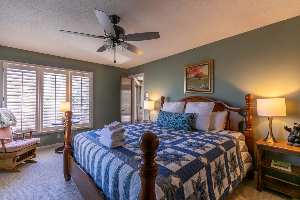 A welcoming bedroom with a soft quilted bed, window with classic shutters, bedside lamps for reading, and tasteful wall art. Gentle morning light enhances the cozy, peaceful setting.