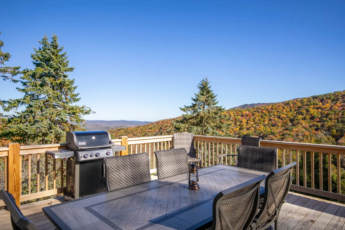 A cheerful outdoor dining area on the deck featuring a table with chairs and a grill, all set against a backdrop of rolling green mountains and towering trees. The scene captures a lively, inviting atmosphere, ideal for barbecues and family gatherings.