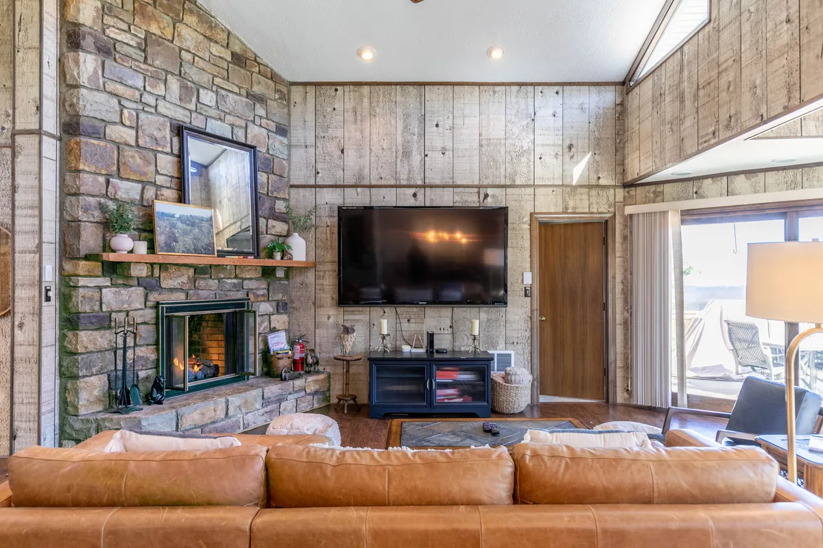 The living room features a prominent stone fireplace, a large television, a comfortable brown leather sofa, wooden wall panels, and a generous window framing outdoor views. The overall mood is warm and inviting, perfect for chilly mountain nights.