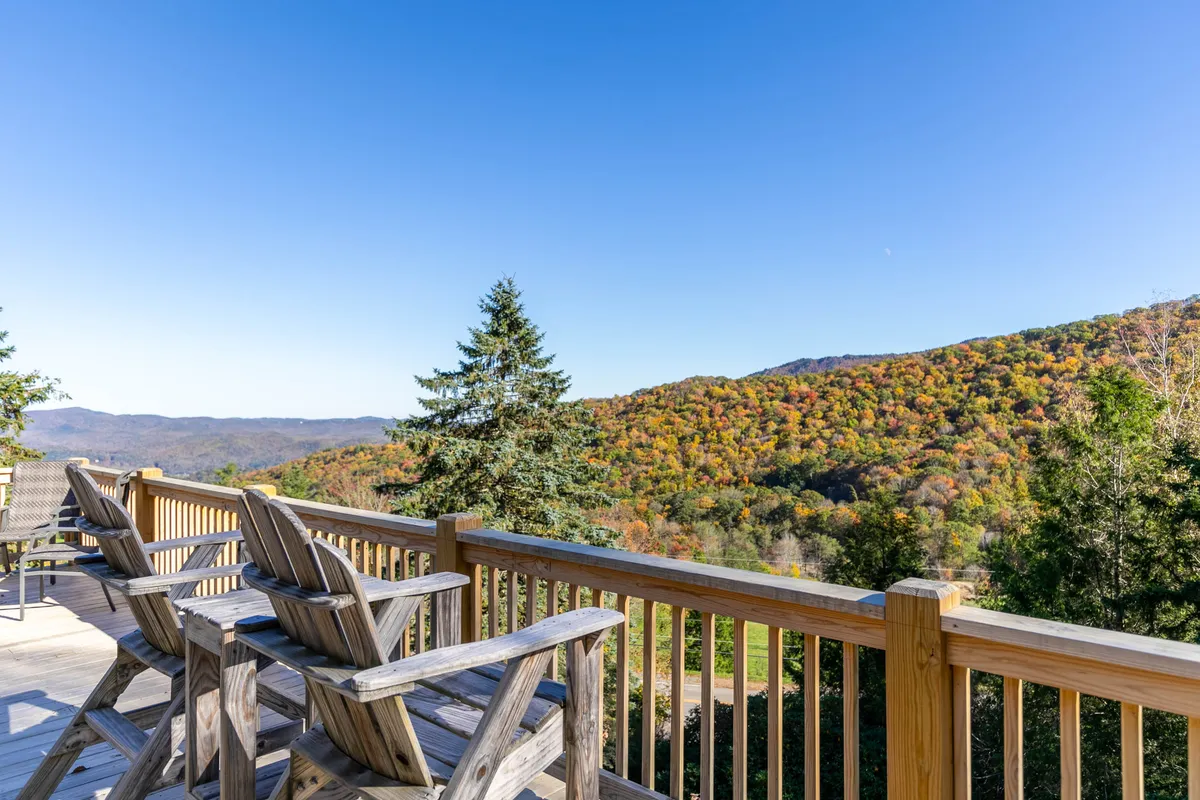 A spacious wooden deck with Adirondack chairs facing out toward a sweeping mountain view. The deck is surrounded by lush trees and vibrant fall foliage, creating a tranquil setting for relaxation and socializing. The expansive deck area is perfect for gatherings or quiet moments taking in the scenery.